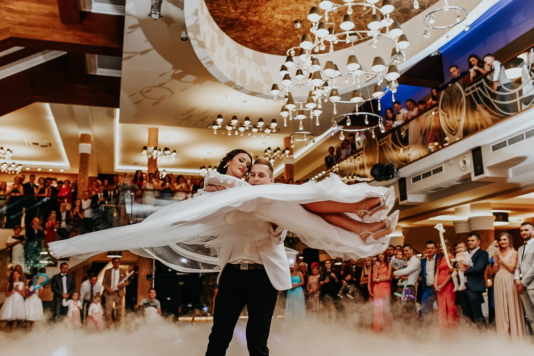 para mloda w dynamicznym tancu slubnym z falujaca suknia trzymana na rekach mlodego pana w nowoczesnej sali slubnej w otoczeniu gosci weselnych patrzacyhc z podziwem na wirujace sylwetki. fotograf grygierczyk lukasz .
