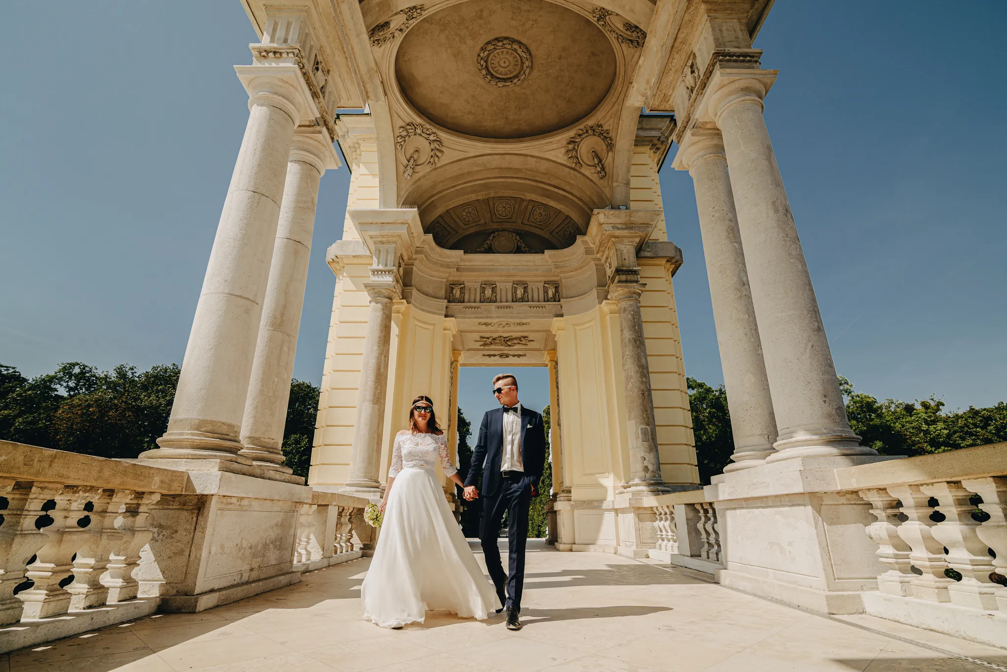 para mloda spaceruje w museum glorietta w pieknym architektonicznie budynku trzymajac sie za rece .pani mloda ma w zwiewnej sukience z wiankiem na glowie a pan mlody w granatowym garniturze. oboje malzonkow posiada zalozone okulary sloneczne