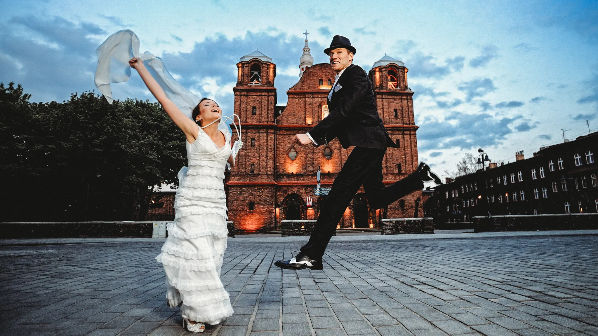 dynamiczny wyskok mlodej pary podczas sesji slubnej w zabytkowej czesci miasta stylizowany na lata dwudzieste dwudziestego wieku . Pan mlody ubrany w stylu al capone z kapeluszem i czarno bialymi butami a pani mloda w stylizowanej fryzurze koralamina szyji na tle Kościół świętej Anny
w Katowicach