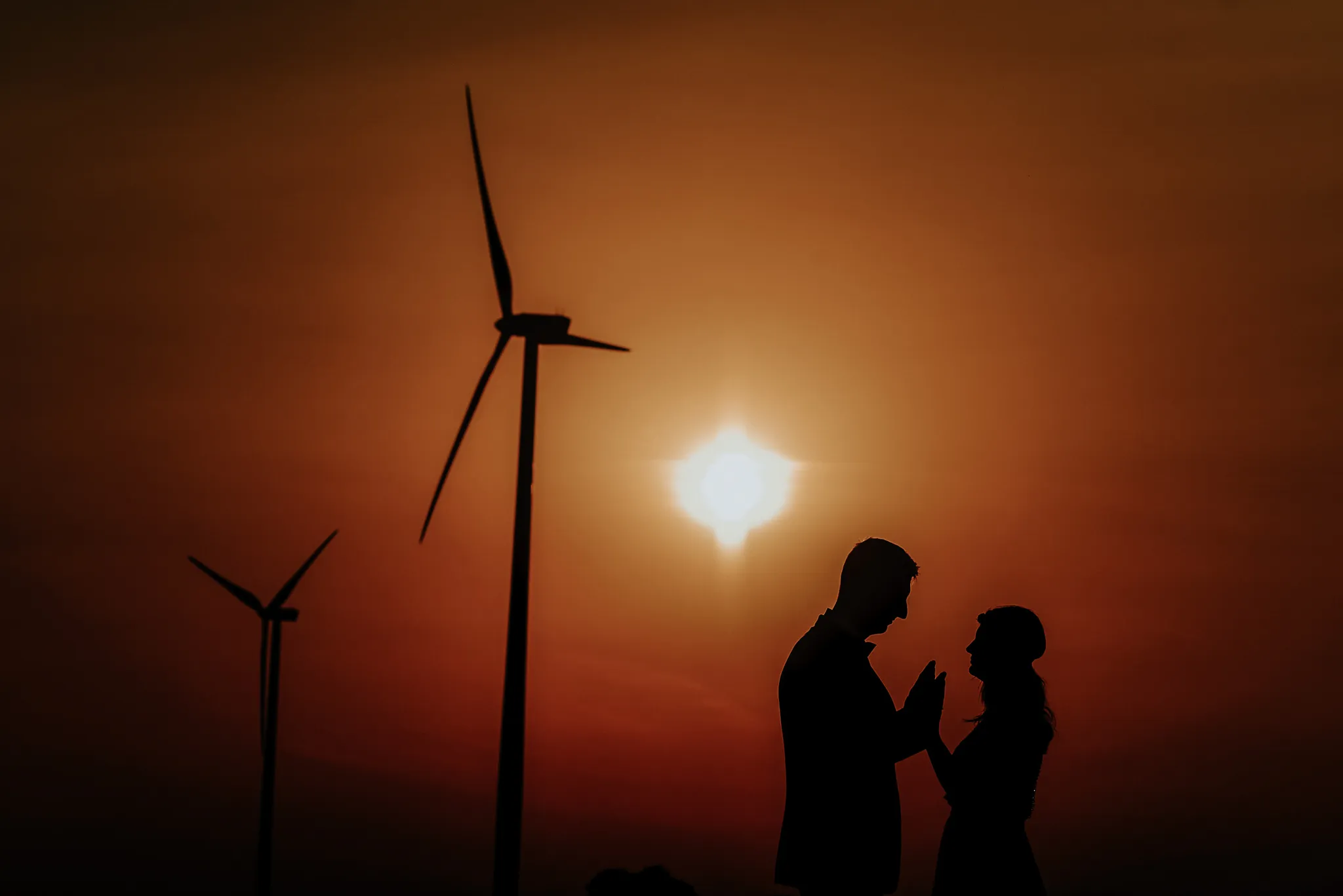 para mloda na tle wiatrakow o zachodzie slonca zwocona do siebie twarzami i trzymajaca sie za rece. w tle zachodzace slonce z pomaranczowa poswiata.