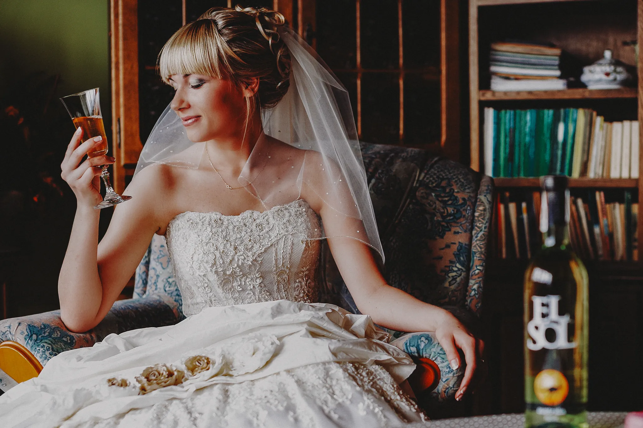 pani mloda siedzaca na fotelu przed ceremonia oczekujaca pana mlodego ubrana w koronkowa biala sukienke delektuje sie lampka wina w stylowym pokoju gdzie jest polka z ksiazkami